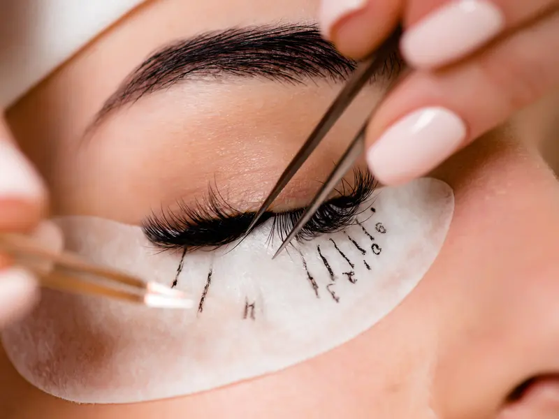 A lash artist isolating one natural lash with two tweezers over an eye pad marked with a lash map